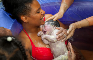 A happy African American mother holding her brand new baby girl after delivering her in a birthing pool at home. The baby is covered in vernix and the mom is laughing.